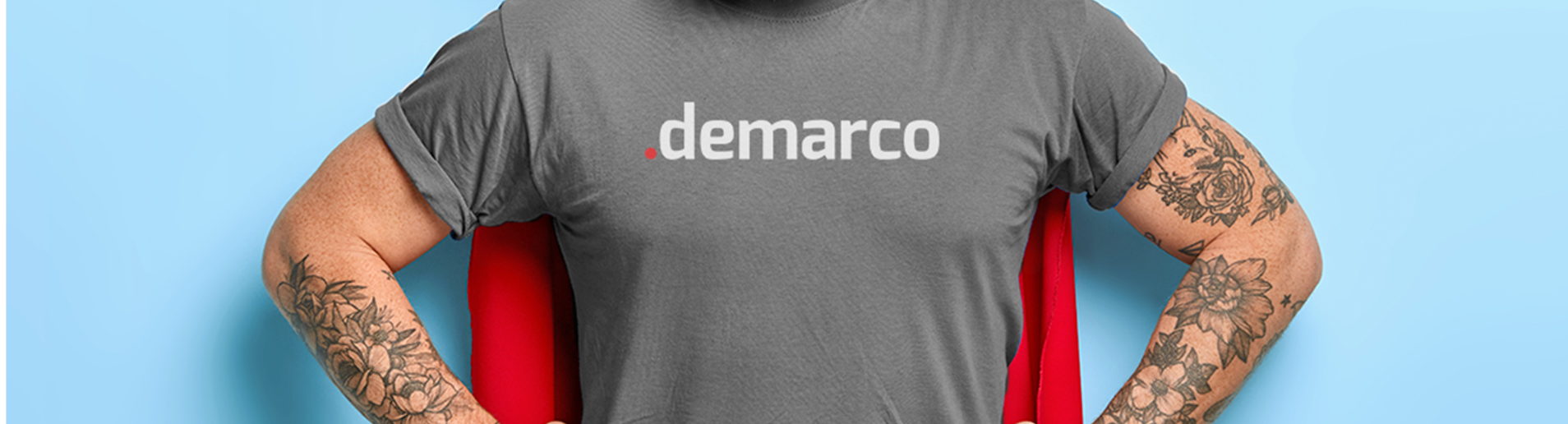 Homem com braços tatuados exibindo uma camiseta cinza com o logotipo 'demarco' em letras brancas, sendo o ponto inicial em vermelho. Ele está com os braços dobrados, destacando as tatuagens de flores e outros detalhes nos antebraços. Ao fundo, há uma parede azul clara, e a cadeira vermelha adiciona um toque de cor vibrante à composição. A imagem transmite uma sensação de atitude e estilo moderno.