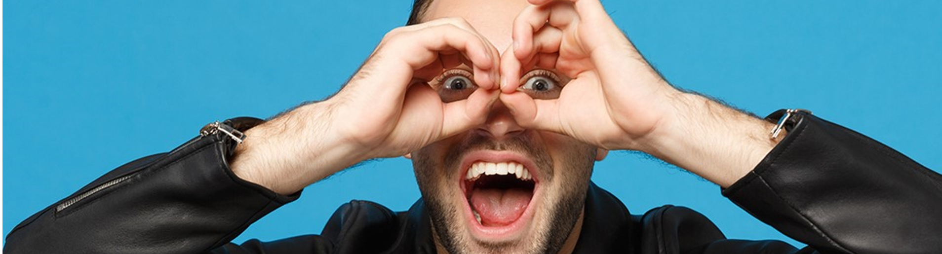 Homem sorridente fazendo um gesto divertido com as mãos em forma de círculo ao redor dos olhos, simulando binóculos. Ele está usando uma jaqueta de couro preta e expressa entusiasmo com a boca aberta. O fundo é azul vibrante, destacando ainda mais sua expressão lúdica e energética.